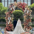 A picture of a white floral arch background with garlands and bouquets. The floral arch is decorated with white roses, daisies, carnations and other flowers. The flower arch is located on a green lawn with blue sky and white clouds in the background. This image is the perfect backdrop for a wedding or other celebration.