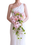 A woman wearing a one-shoulder white wedding dress holds a large cascading bouquet of pink roses, white flowers, and green leaves against a plain white background.