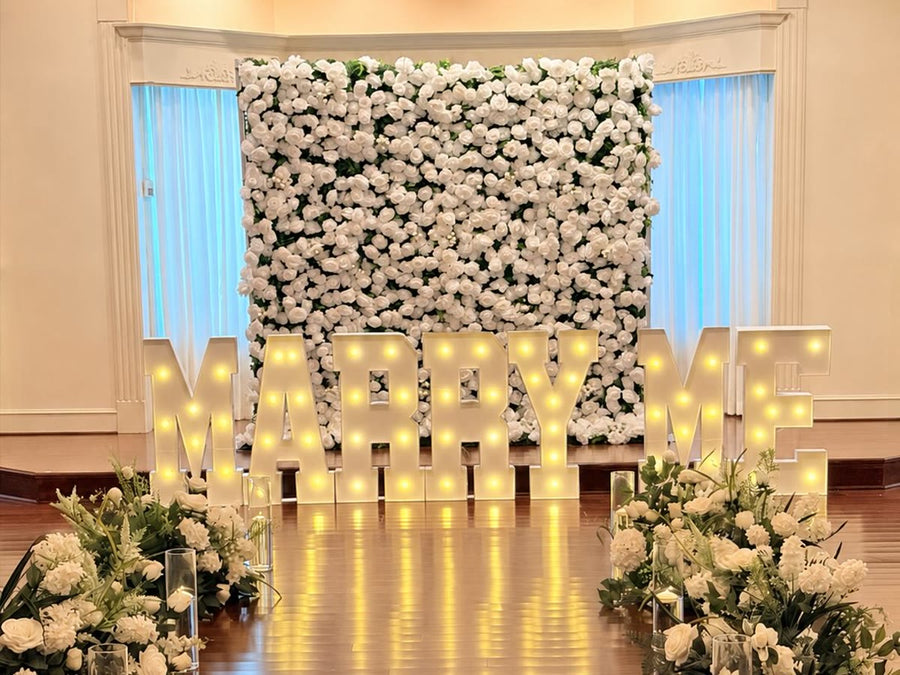 White rose flower wall behind illuminated MARRY ME letters